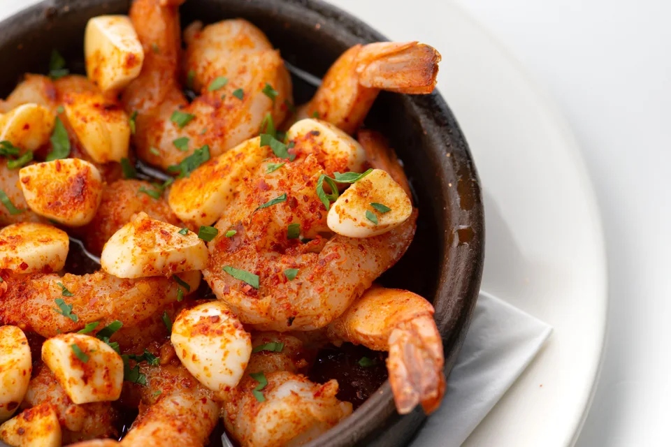 BookNoFees: Foto de ejemplo: Gambas al ajillo adornadas con hierbas picadas en un cuenco negro sobre un plato blanco